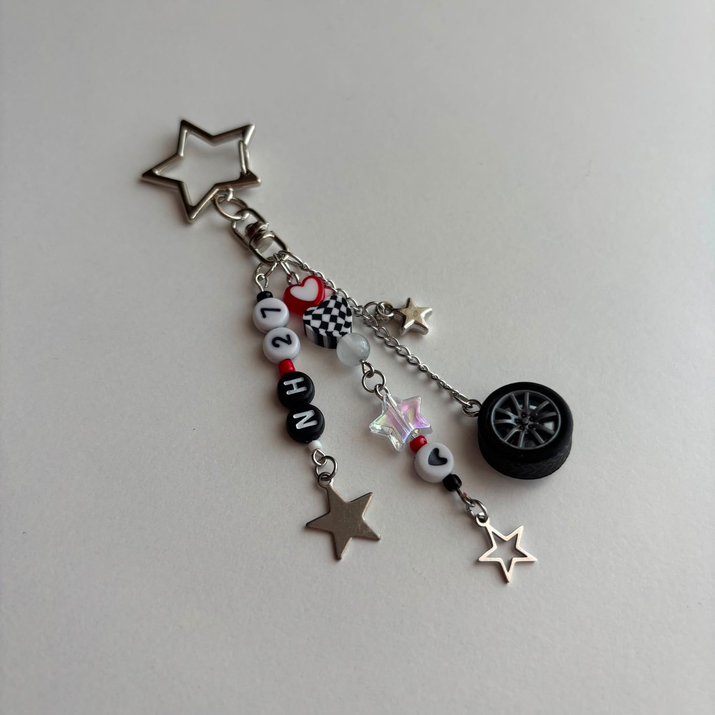 A silver and black car-themed charm keychain featuring a star-shaped keyring, letter beads spelling 'NH27', a checkered flag charm, a small heart charm, a miniature tire, and star charms on a light gray background.