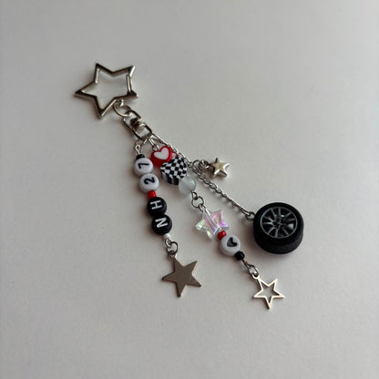 A silver and black car-themed charm keychain featuring a star-shaped keyring, letter beads spelling 'NH27', a checkered flag charm, a small heart charm, a miniature tire, and star charms on a light gray background.
