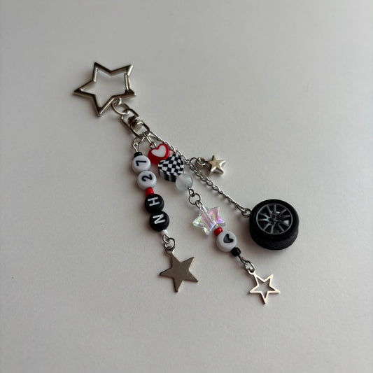 A silver and black car-themed charm keychain featuring a star-shaped keyring, letter beads spelling 'NH27', a checkered flag charm, a small heart charm, a miniature tire, and star charms on a light gray background.