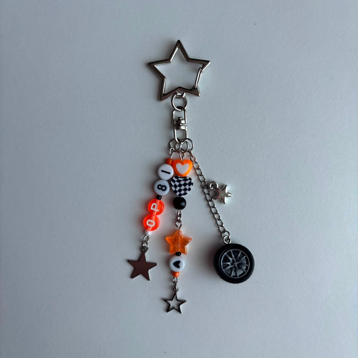 A silver metal keychain charm featuring a star-shaped top, a small heart, and dangling charms including orange and black beads with a checkered heart, a mini star, a brown star, and a small tire charm, displayed on a light gray background