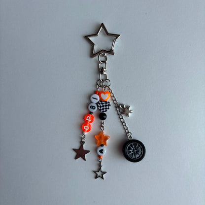 A silver metal keychain charm featuring a star-shaped top, a small heart, and dangling charms including orange and black beads with a checkered heart, a mini star, a brown star, and a small tire charm, displayed on a light gray background