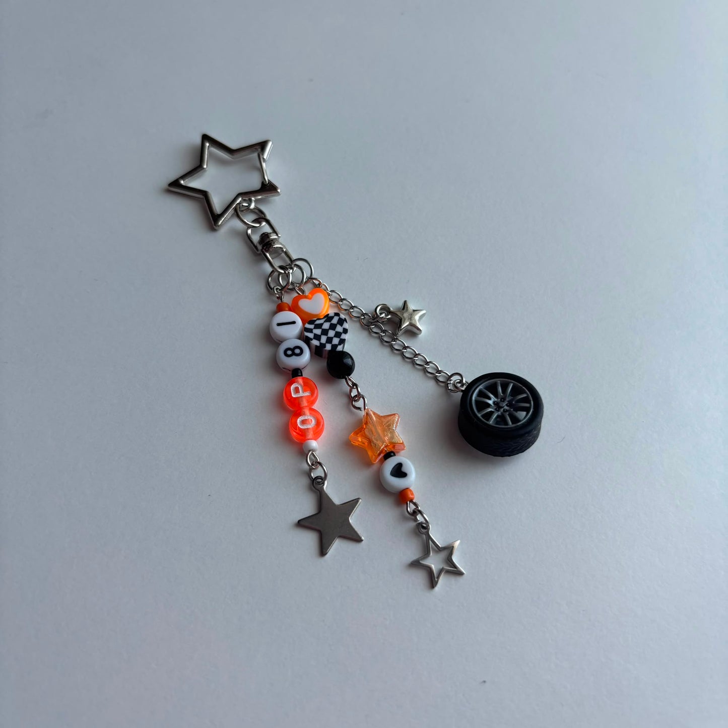A silver-tone metal charm keychain featuring a star-shaped keyring, a checkered flag charm, orange and white bead accents, a small black tire charm, and two dangling star charms on a light gray background.