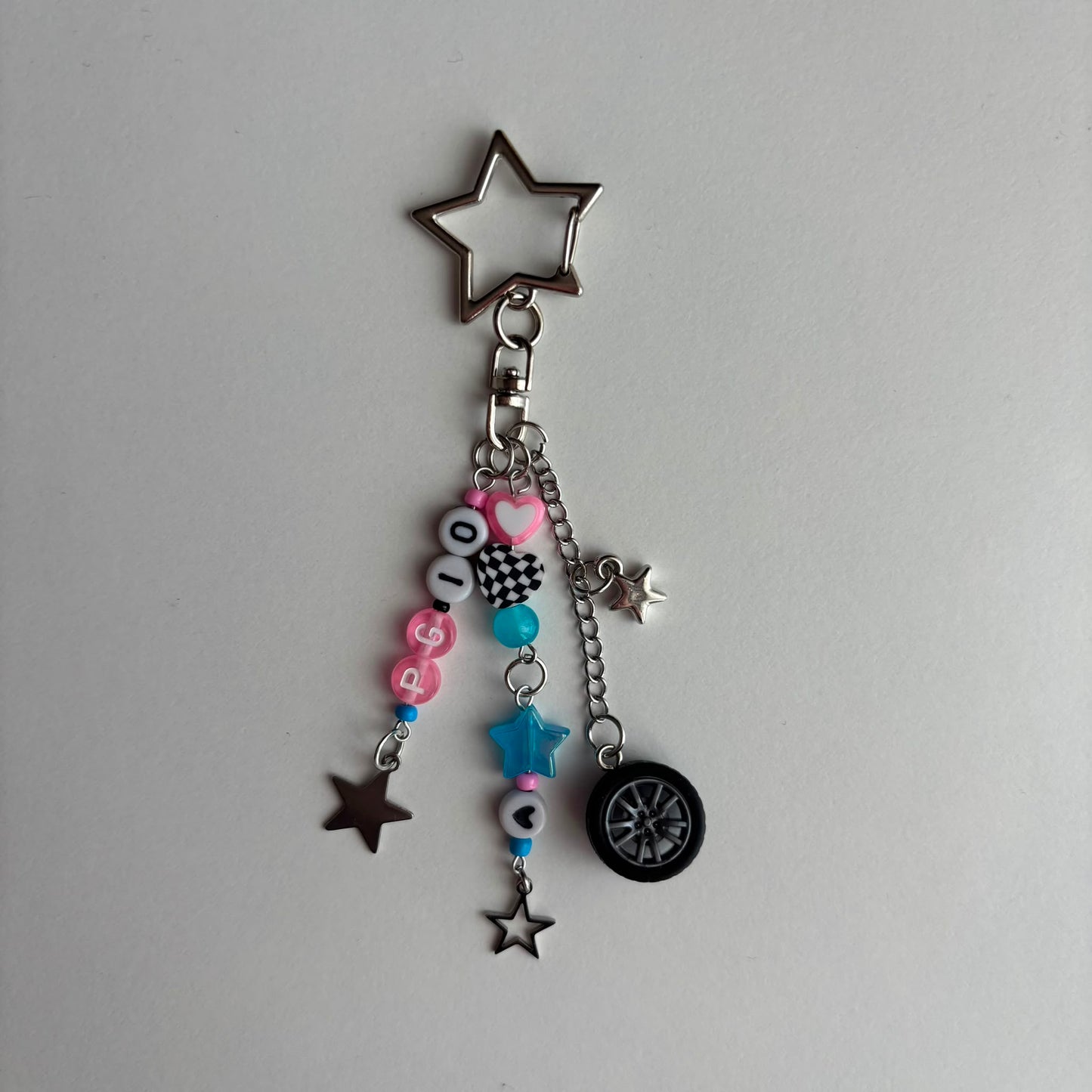 A silver metal keychain charm featuring a large star-shaped keyring, a clasp, and dangling charms including pink and black letter beads spelling 'PG10', a blue bead, a small star, a mini tire, and several smaller star charms, displayed on a plain light gray background.