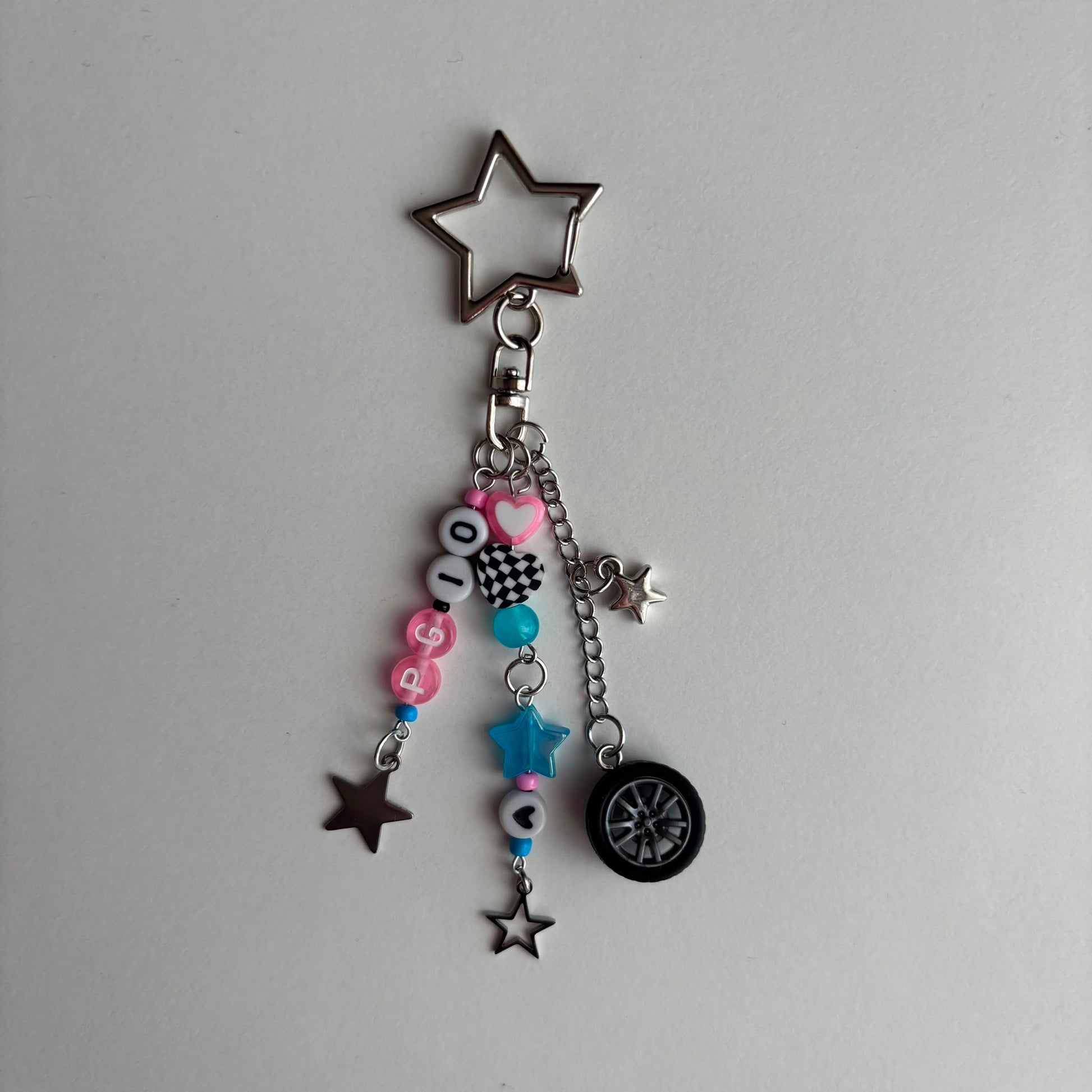 A silver metal keychain charm featuring a large star-shaped keyring, a clasp, and dangling charms including pink and black letter beads spelling 'PG10', a blue bead, a small star, a mini tire, and several smaller star charms, displayed on a plain light gray background.