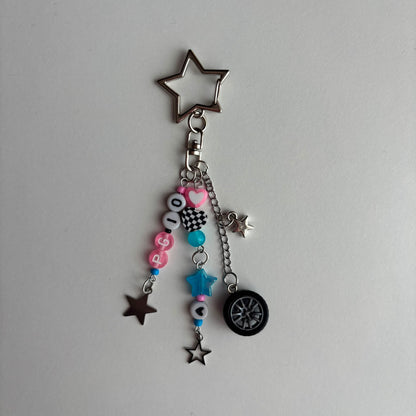 A silver metal keychain charm featuring a large star-shaped keyring, a clasp, and dangling charms including pink and black letter beads spelling 'PG10', a blue bead, a small star, a mini tire, and several smaller star charms, displayed on a plain light gray background.