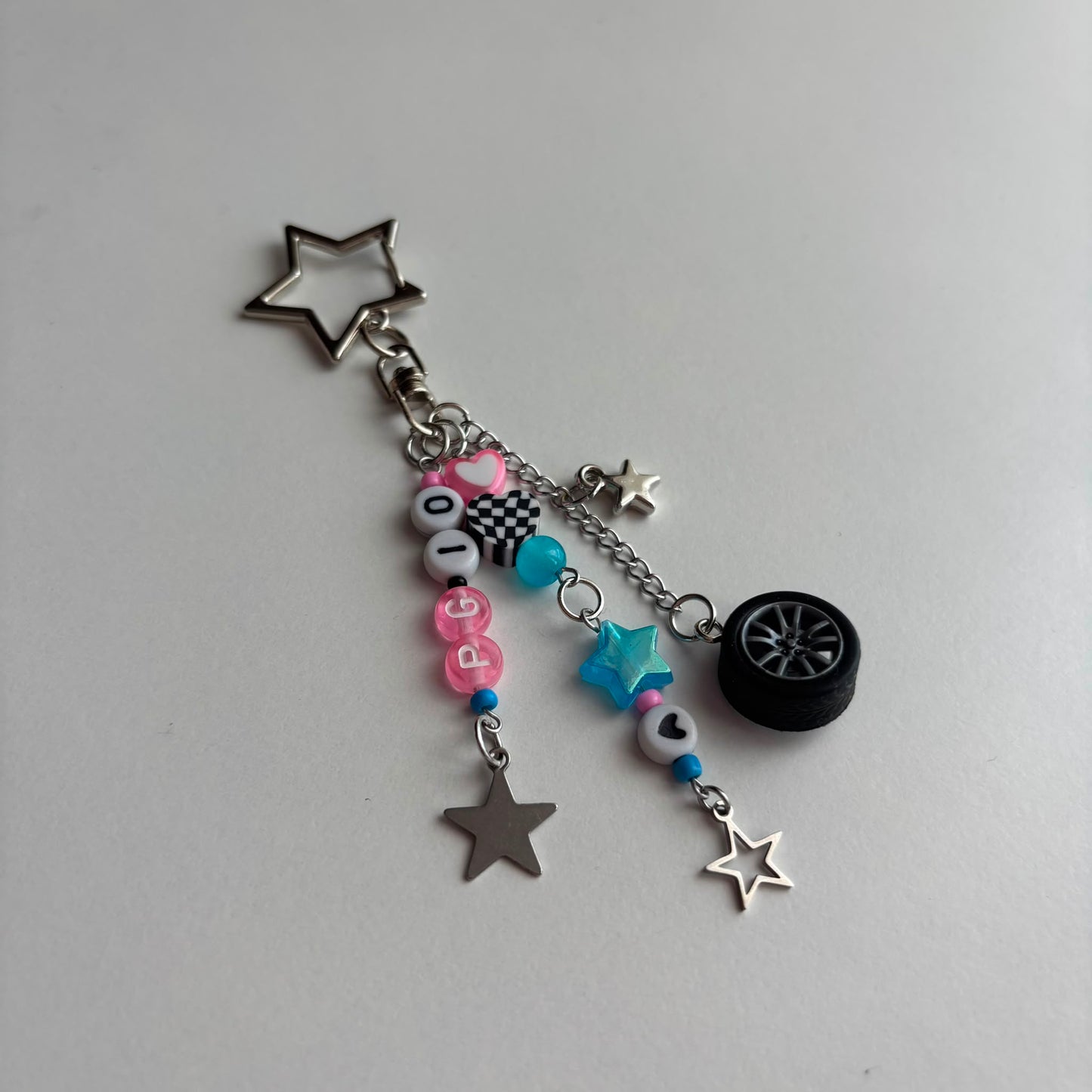 A silver-toned keychain charm featuring a star-shaped ring, a small tire charm, and dangling beaded chains with pink, blue, and white beads, silver star charms, and a checkered bead.