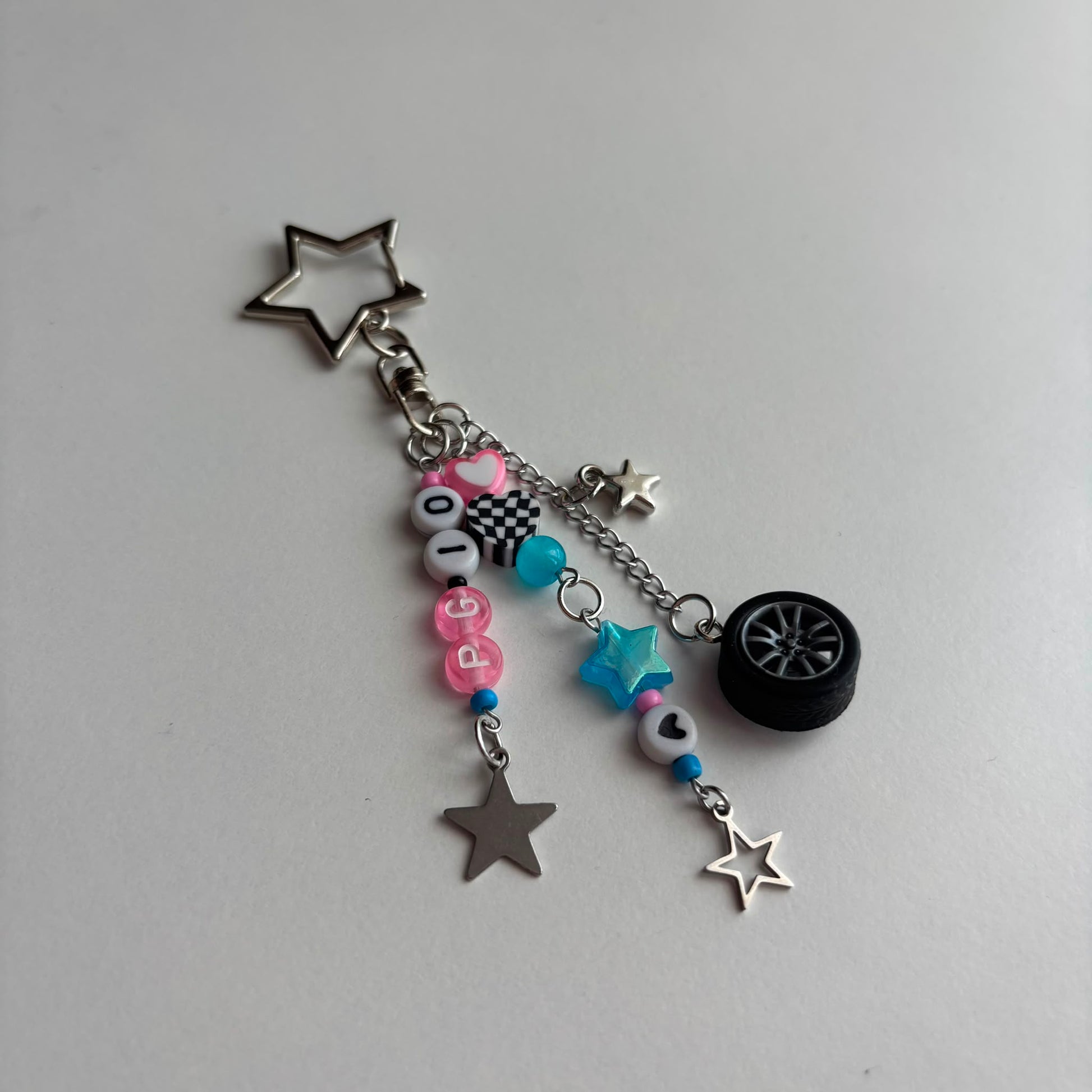 A silver-toned keychain charm featuring a star-shaped ring, a small tire charm, and dangling beaded chains with pink, blue, and white beads, silver star charms, and a checkered bead.