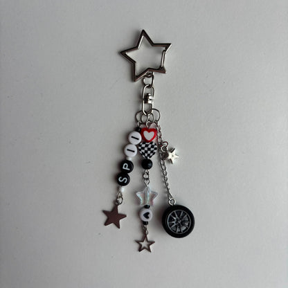 A silver metal keychain charm featuring a star-shaped top, a split ring, and multiple dangling charms including beads spelling 'SP11', a red heart, checkered squares, star charms, a small silver star, and a black-and-silver tire charm, all photographed on a plain light background.