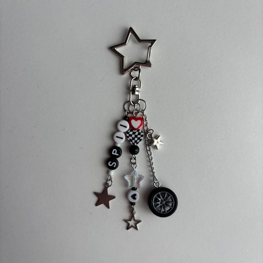 A silver metal keychain charm featuring a star-shaped top, a split ring, and multiple dangling charms including beads spelling 'SP11', a red heart, checkered squares, star charms, a small silver star, and a black-and-silver tire charm, all photographed on a plain light background.