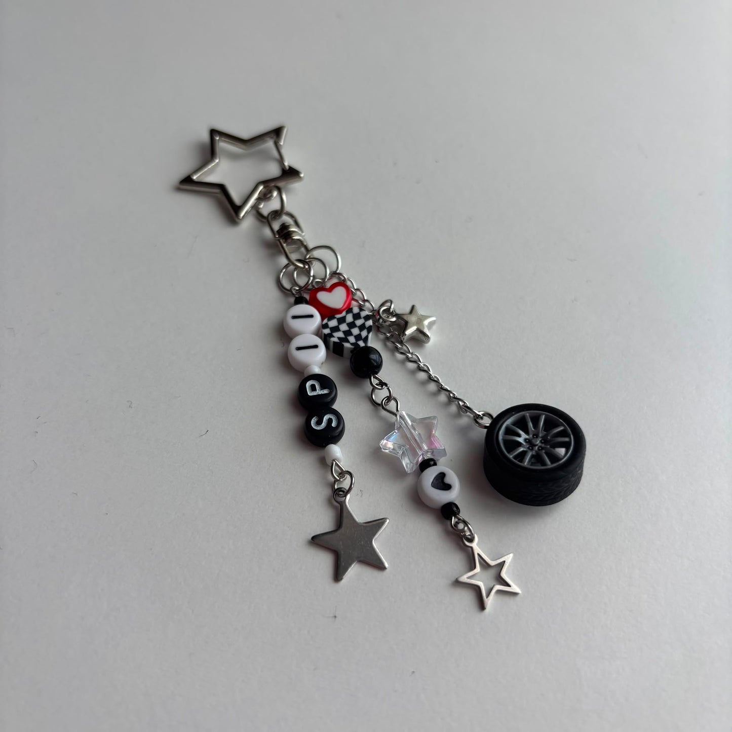 A silver-tone metal keychain charm featuring star-shaped pendants, a miniature black car wheel, black and white beads spelling 'SP11', a checkered heart, and a small red heart accent, displayed on a light gray background.