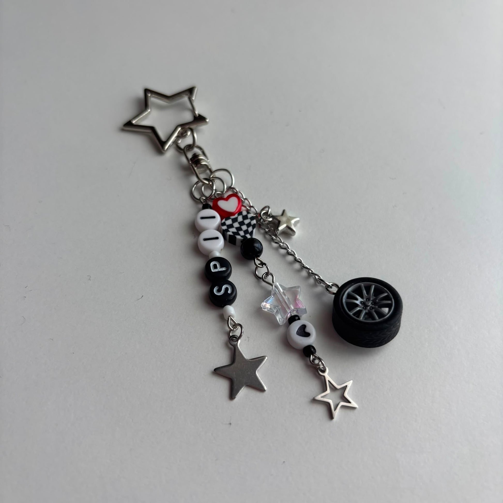 A silver-tone metal keychain charm featuring star-shaped pendants, a miniature black car wheel, black and white beads spelling 'SP11', a checkered heart, and a small red heart accent, displayed on a light gray background.