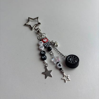A silver-tone metal keychain charm featuring star-shaped pendants, a miniature black car wheel, black and white beads spelling 'SP11', a checkered heart, and a small red heart accent, displayed on a light gray background.