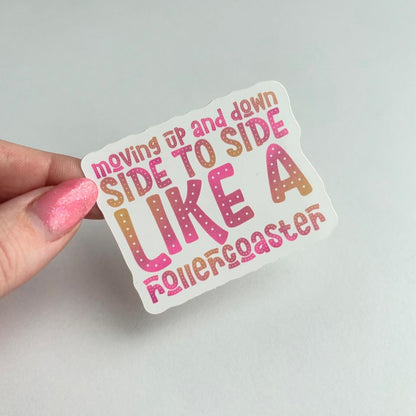 A hand with pink glitter nail polish holds a die-cut sticker that reads 'moving up and down SIDE TO SIDE LIKE A rollercoaster' in pink, orange, and brown gradient lettering on a white background, against a light gray wall.