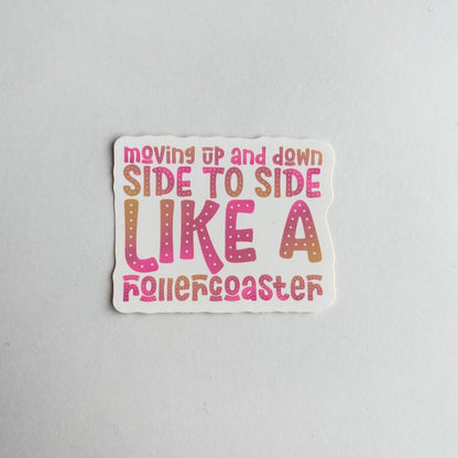 A die-cut sticker with the phrase 'moving up and down SIDE TO SIDE LIKE A rollercoaster' in pink, orange, and brown gradient lettering on a white background, photographed on a light gray surface.